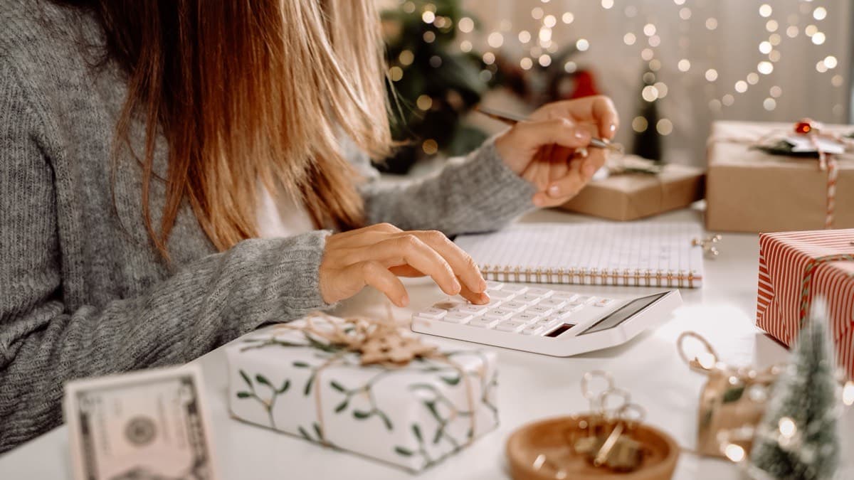 A woman working out holiday costs on a calculator and notebook among wrapped gifts and cash