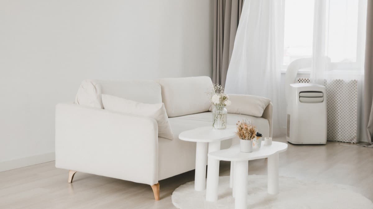A bright apartment living room with a white sofa and a portable air conditioner by the window