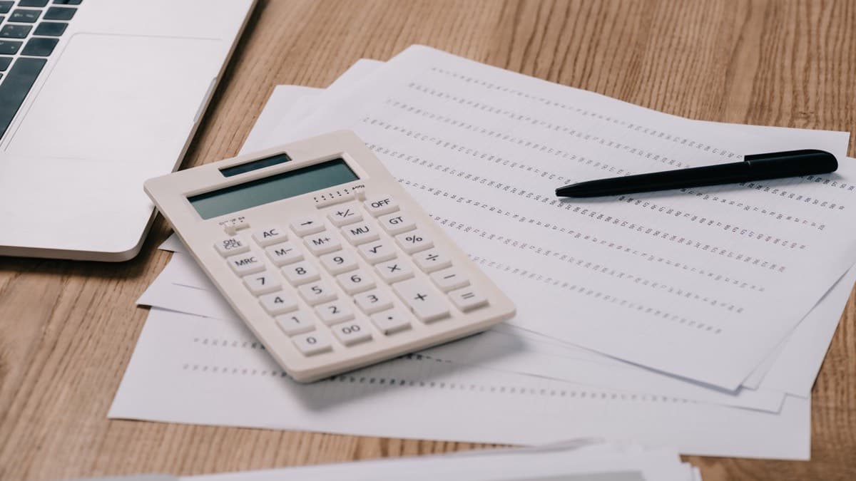 A calculator and a pen on a spread of printed pages beside a laptop on a wooden table