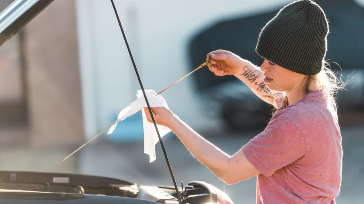 A woman in a beanie reading the oil dipstick with a rag under an open car hood