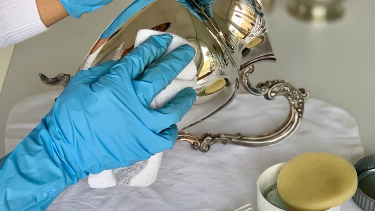 Gloved hands polishing a silver pitcher beside a dish of polish and silverware
