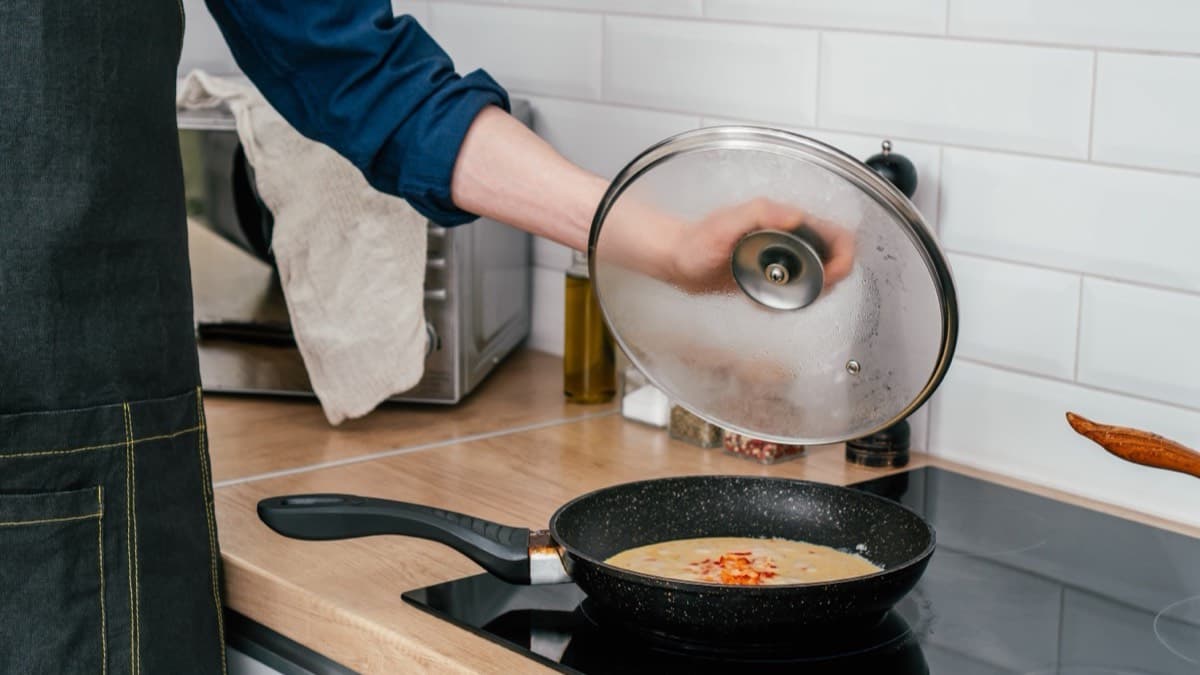A cook lifting a glass lid off a frying pan on a black stovetop