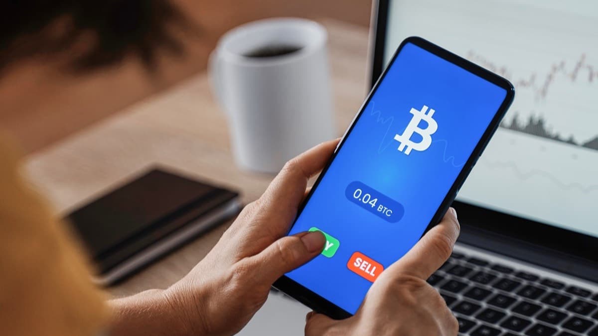 A person holding a phone showing a Bitcoin buy and sell screen at a desk with a laptop