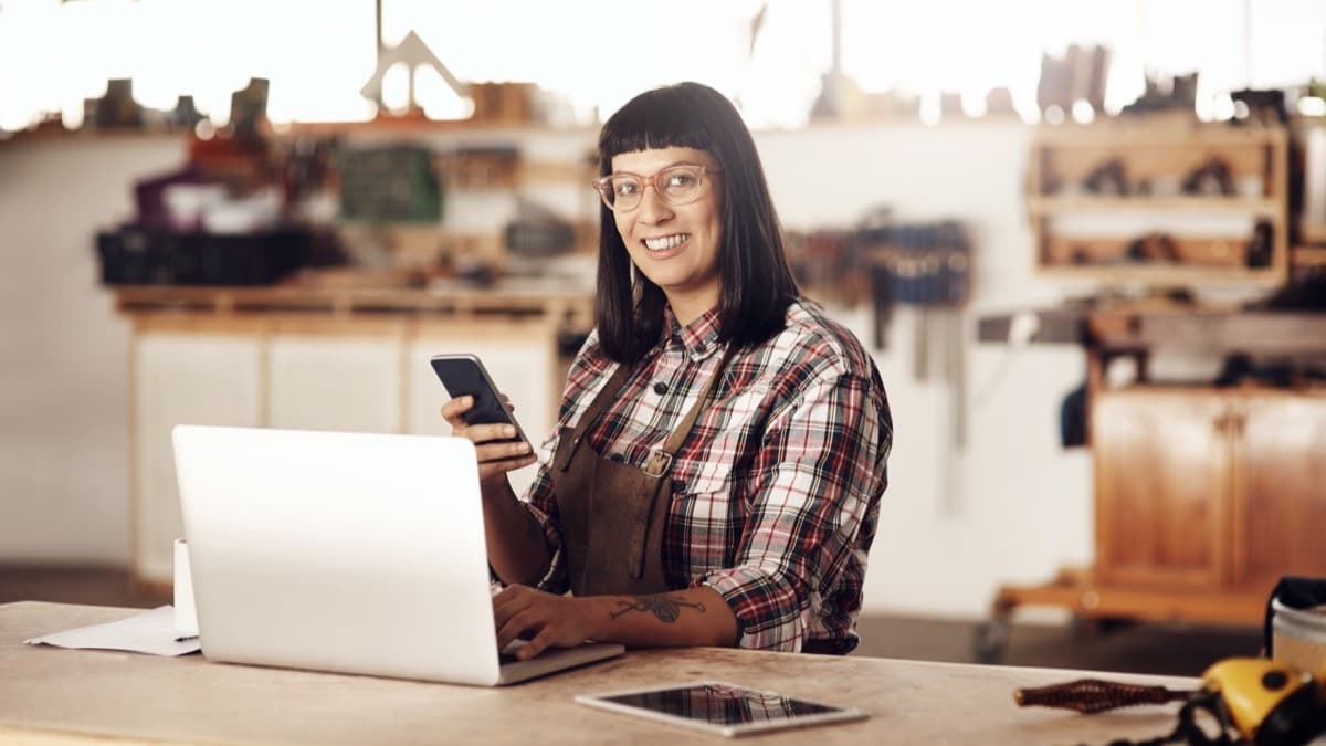 A carpenter at a workshop counter using a laptop with a phone in hand