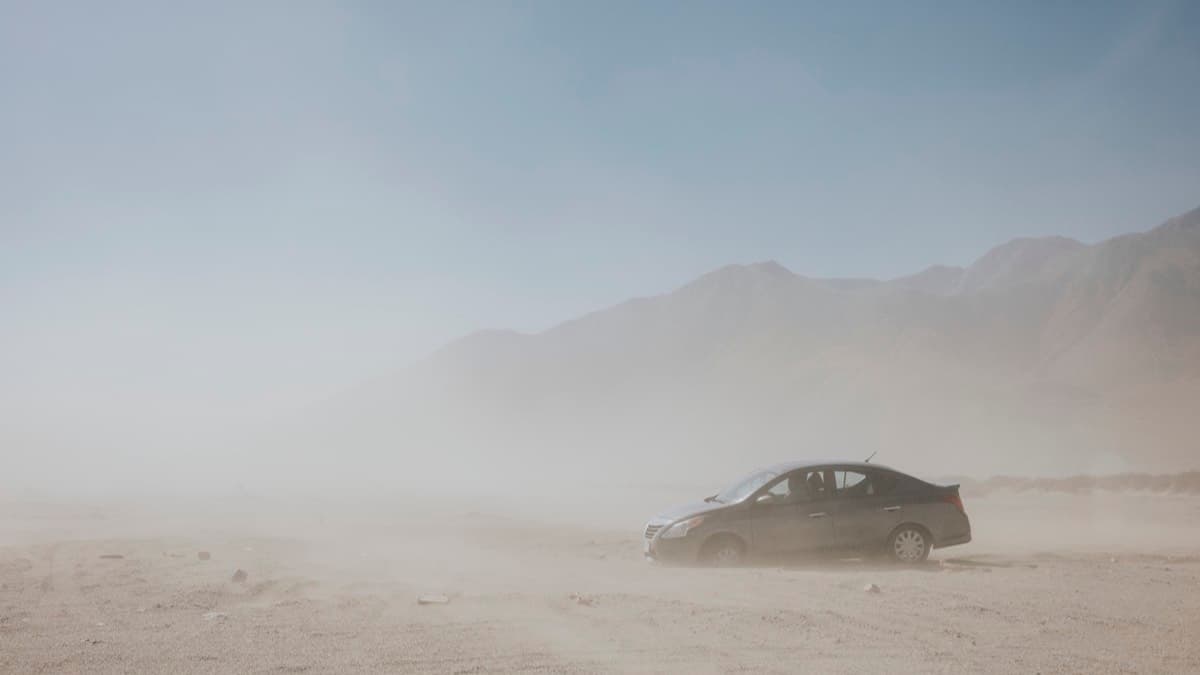 A car stopped off the road in blowing desert dust, mountains faint behind it