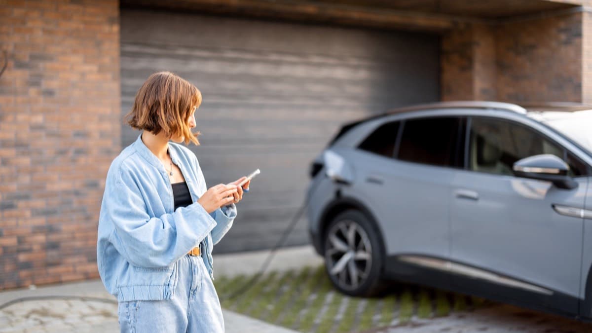 A woman checking her phone in a driveway beside an electric SUV plugged in to charge in front of a garage door
