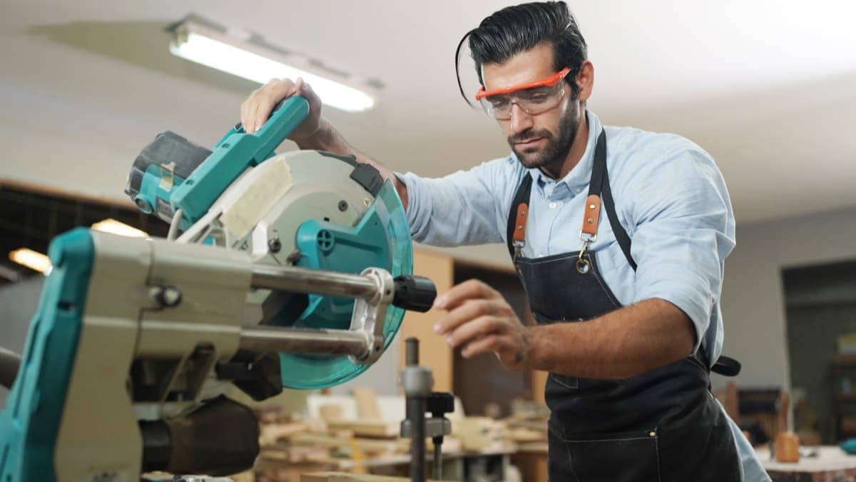 A woodworker in orange-framed safety glasses and a work apron lowering a miter saw onto a board in a workshop