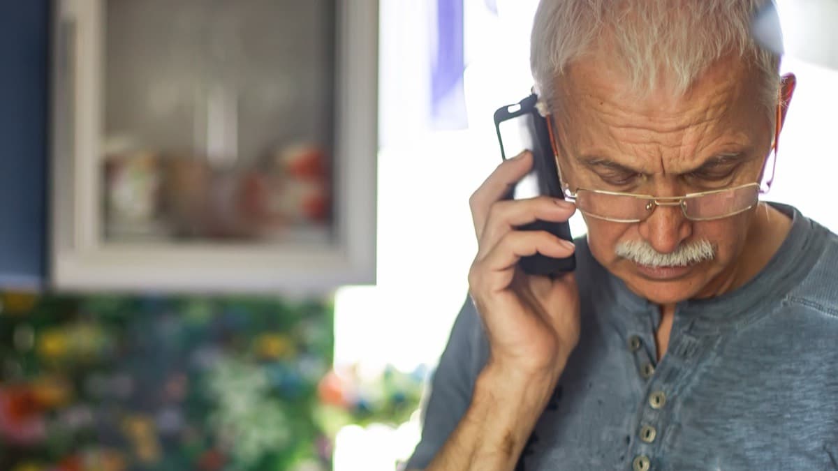 An older man in glasses frowning as he listens on a phone held to his ear at home