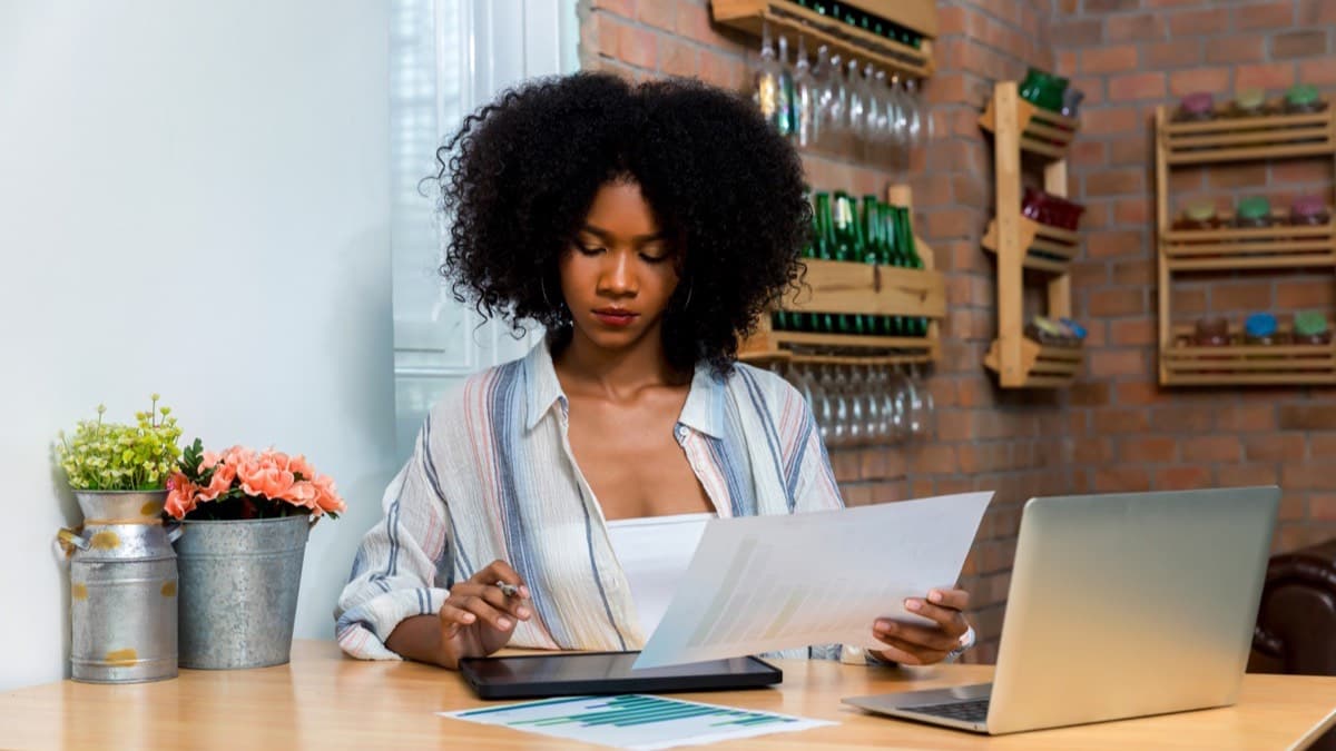 A woman at a desk reading a printed page beside an open laptop and a tablet, a brick wall with shelves behind her