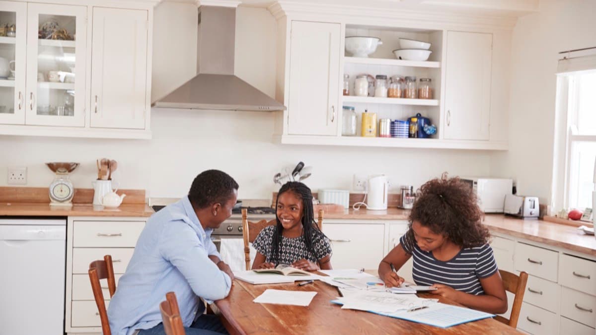 A father and two daughters sitting at a wooden kitchen table spread with papers and notebooks