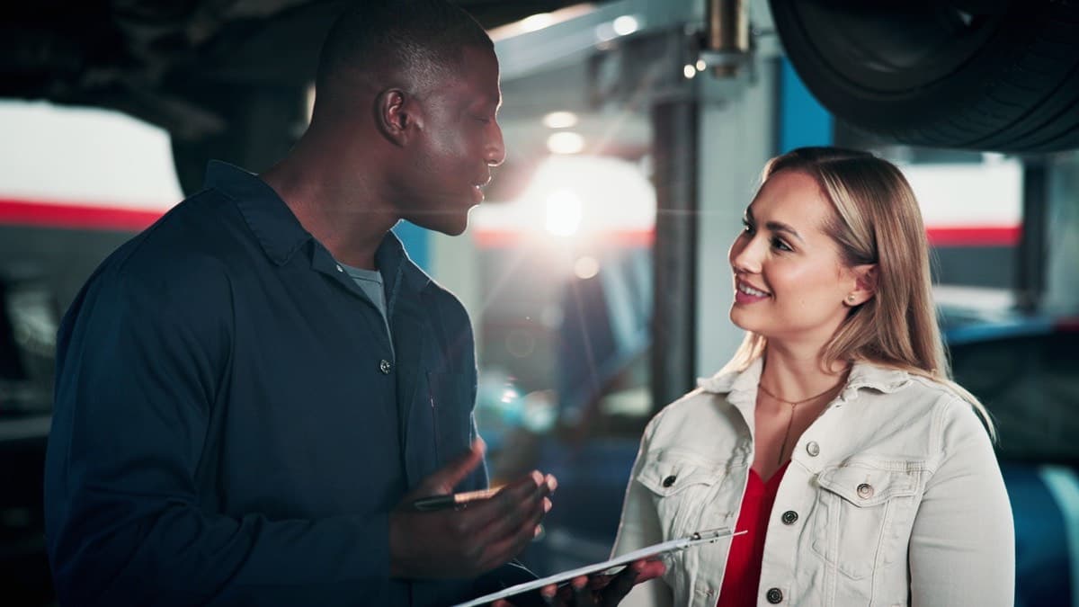A mechanic in coveralls talking with a customer under a raised car in a repair shop