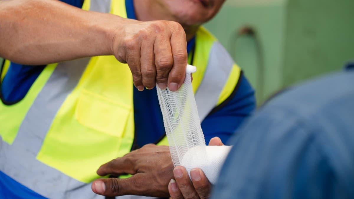 A worker in a hi-vis vest wrapping gauze around a co-worker's injured hand on the job