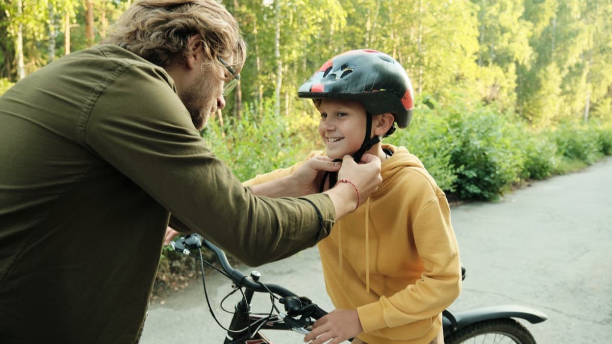 A father fastening the chin strap of his son's bike helmet on a path beside the boy's bicycle