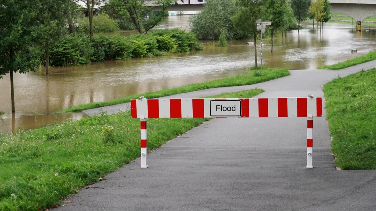 A park path closed by a red and white flood barrier, with a flooded river spreading over the grass beyond it