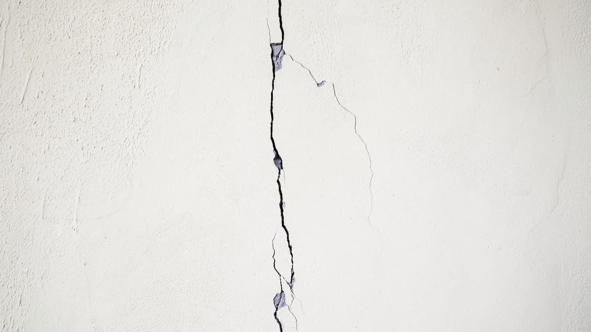 A vertical crack running the full height of a pale plastered wall, with fine hairline cracks branching off it