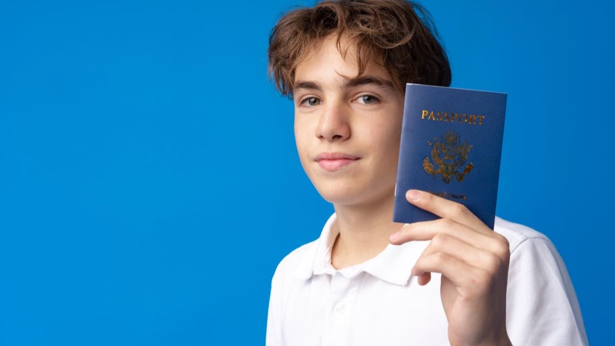 A young teenager holding up a blue United States passport against a bright blue background