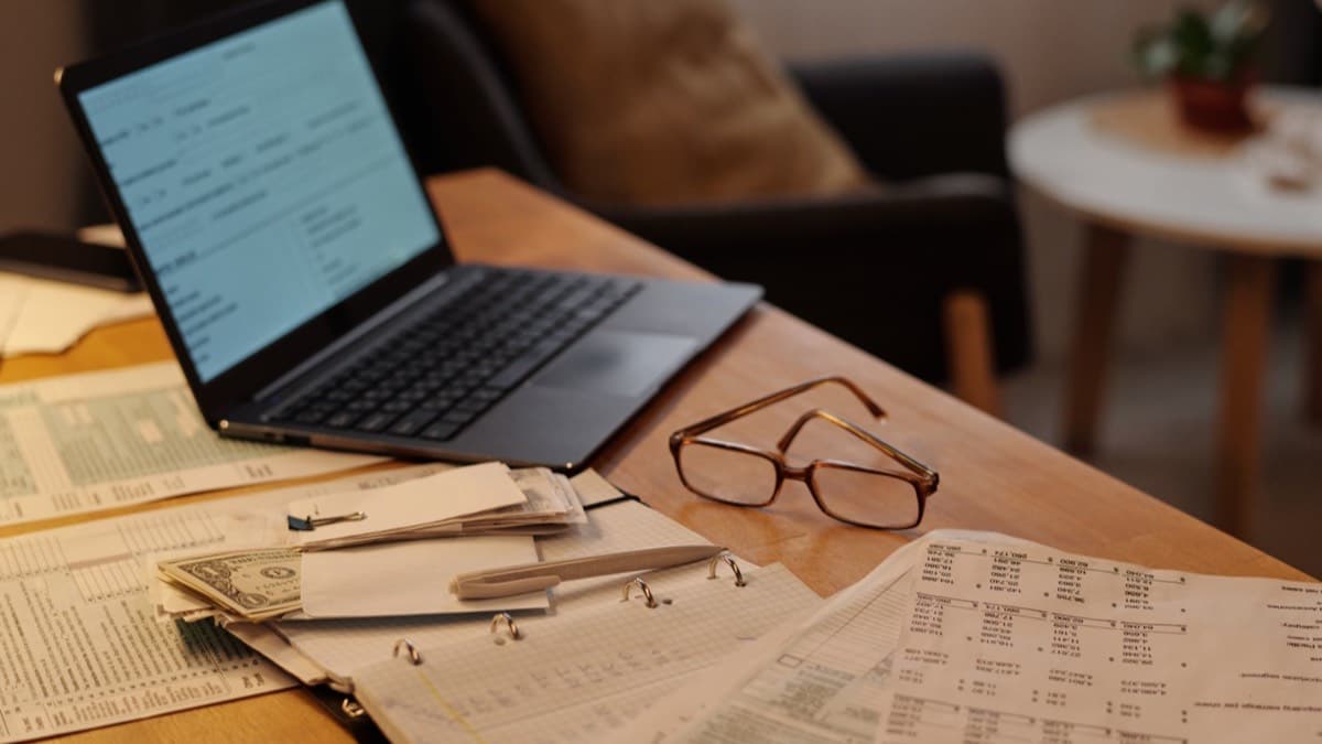 A laptop open beside printed financial forms, a few dollar bills and a pair of reading glasses on a wooden table