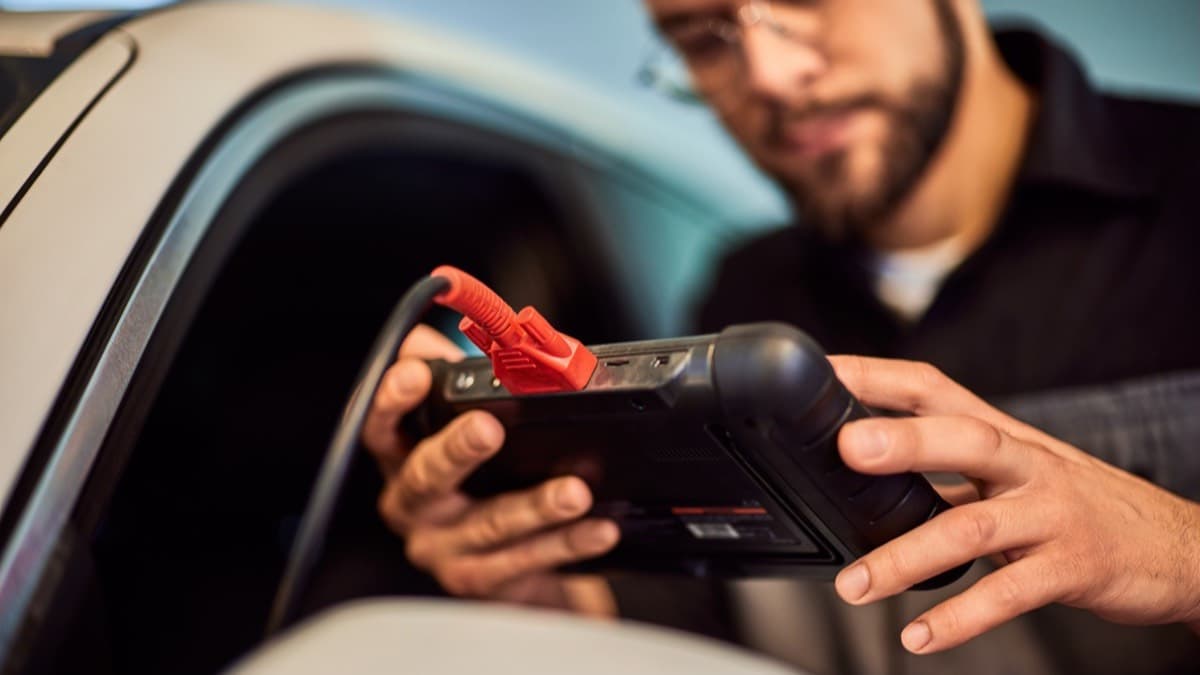 A technician in glasses reading a handheld diagnostic scanner whose cable runs into an open car door