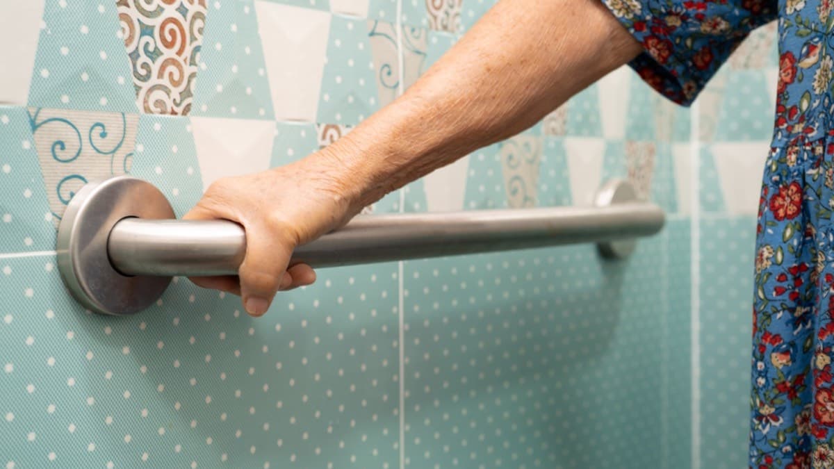 An older woman's hand gripping a stainless steel grab bar mounted on a tiled bathroom wall