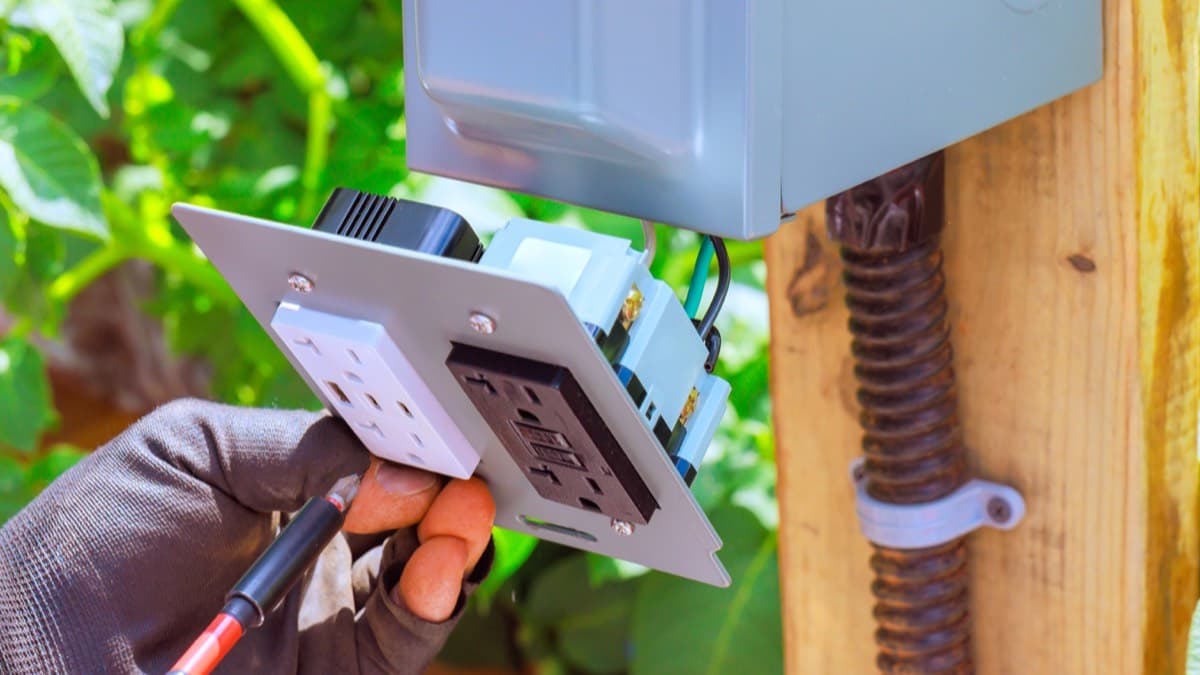 A gloved electrician holding a faceplate with a GFCI outlet, its test and reset buttons between the sockets, beside an outdoor electrical box on a wooden post