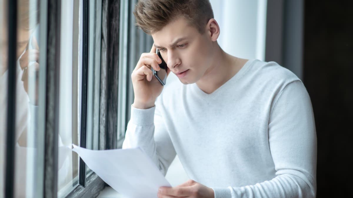 A young man by a window frowning at a letter in his hand while he talks on his phone