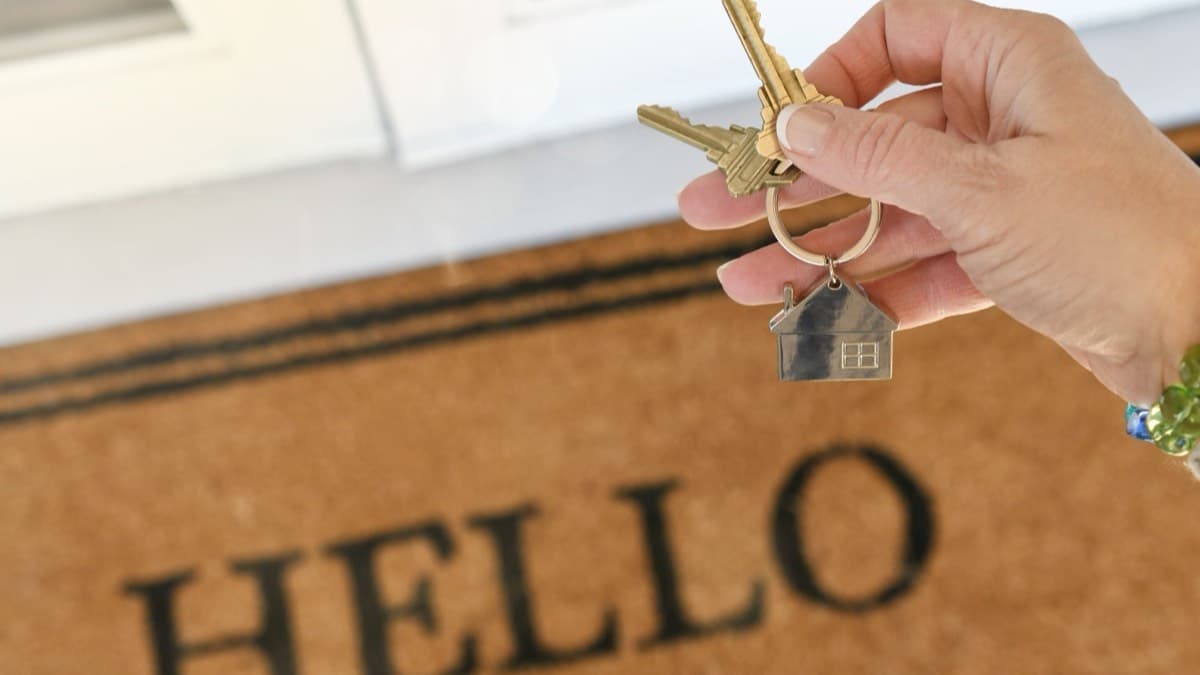 A hand holding a set of house keys on a house-shaped keychain above a HELLO welcome mat at a front door