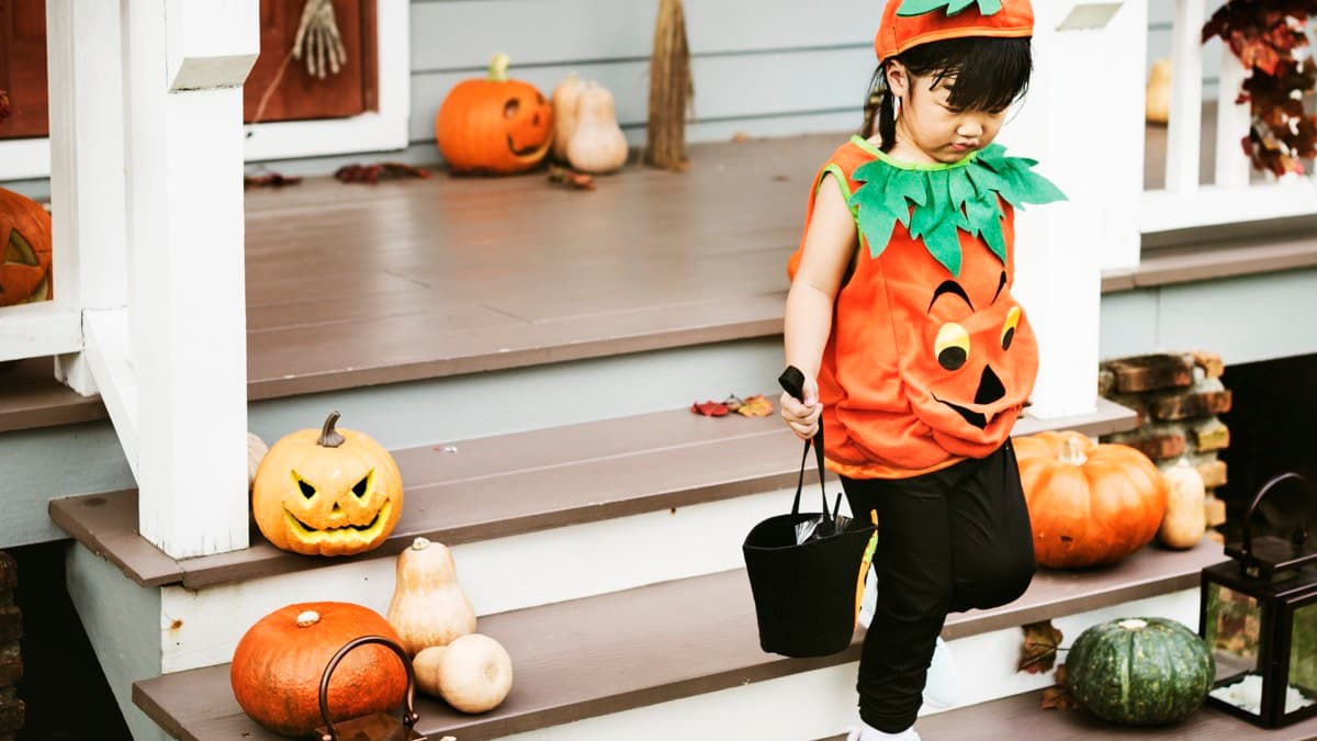 A small child in a pumpkin costume walking down porch steps lined with jack-o'-lanterns, gourds and lanterns