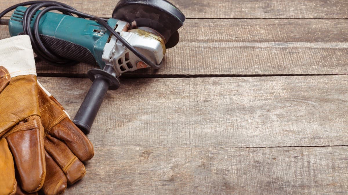 An angle grinder with its cord coiled beside a pair of worn leather work gloves on a weathered wooden workbench