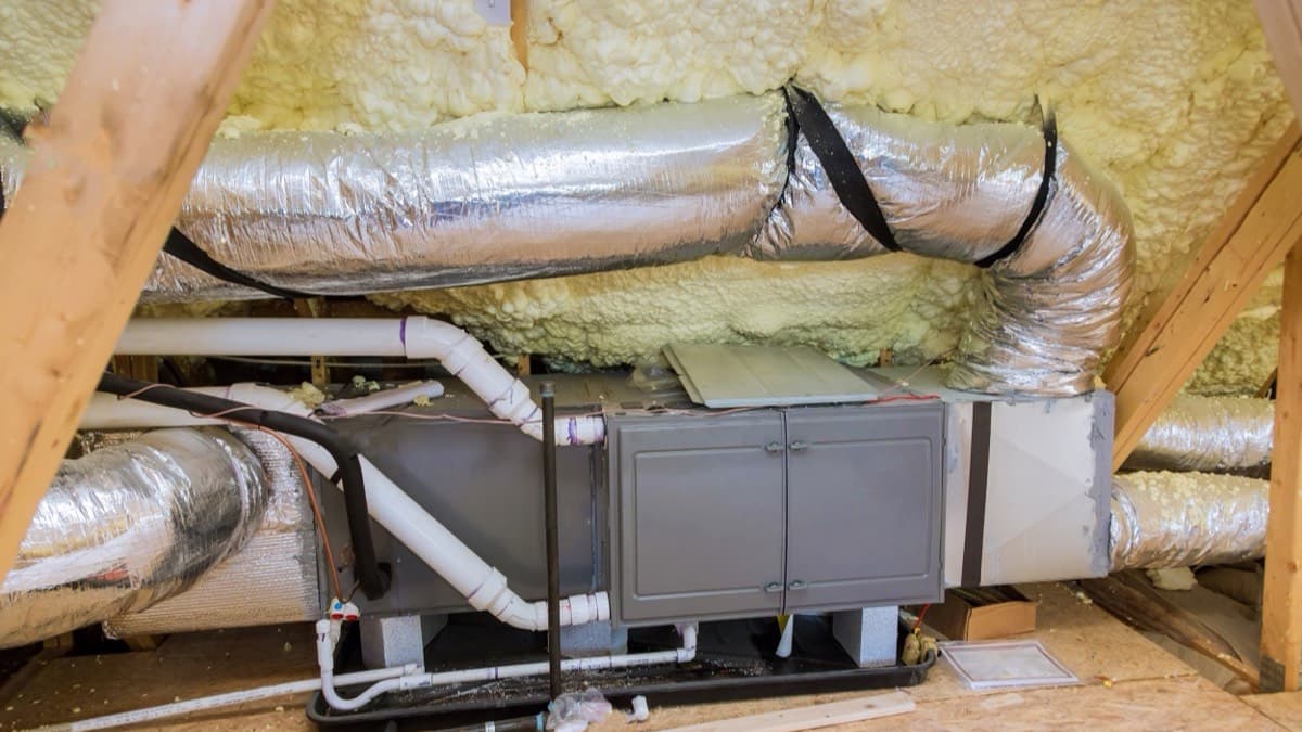A ventilation unit on an attic floor, wrapped insulated ducts running from it under spray-foamed roof rafters
