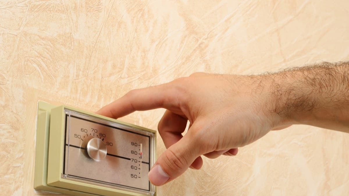 A hand turning down the dial of a beige wall thermostat marked in degrees Fahrenheit