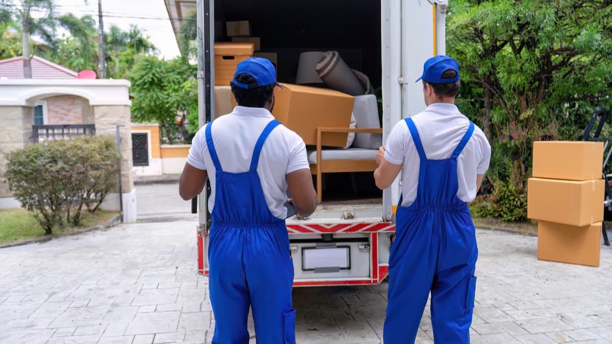 Two movers in blue overalls and caps at the open back of a loaded moving truck in a driveway, boxes stacked beside it