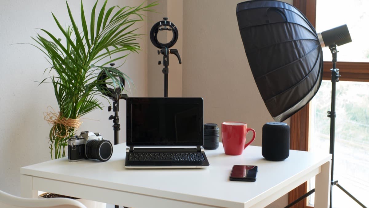A home studio desk with a laptop, a film camera, a speaker and a phone, a ring light and a softbox on stands behind it