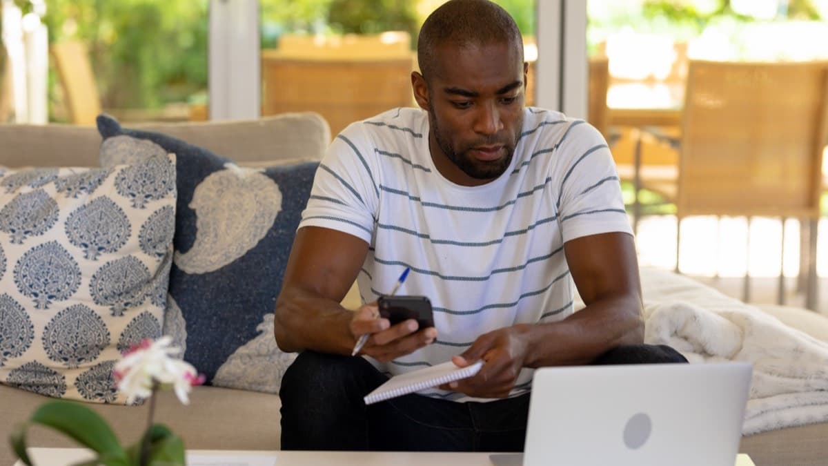 A man on a living room sofa checking his phone with a pen and a sheet of paper in hand, a laptop open on the coffee table