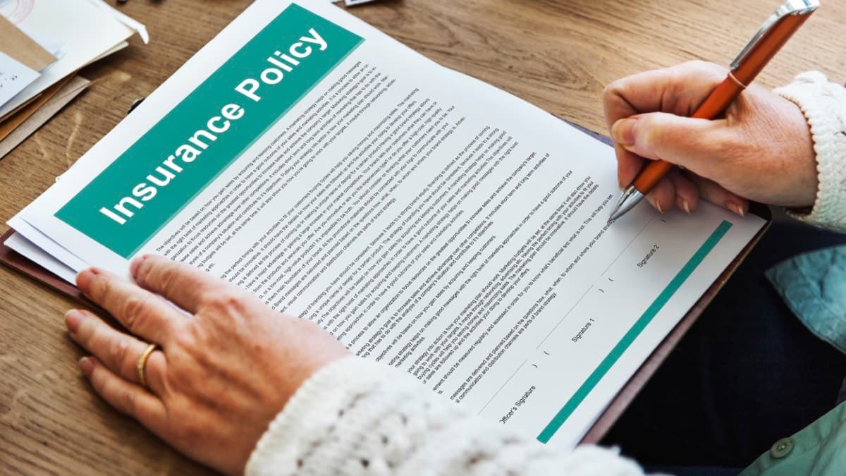 A hand in a knit sweater holding an orange pen over an insurance policy document on a wooden desk