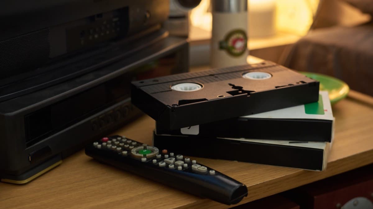 A stack of VHS tapes and a remote on a wooden side table beside an old VCR in warm lamplight