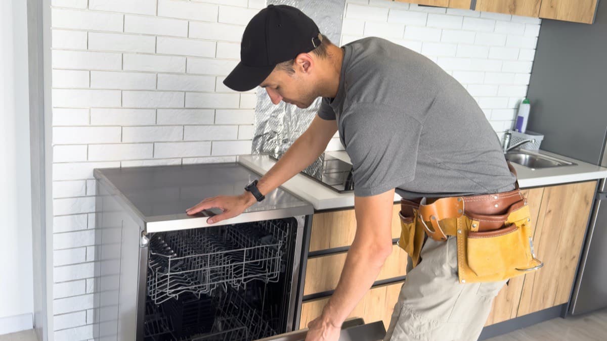 A repair technician in a cap and tool belt leaning over an open dishwasher in a home kitchen