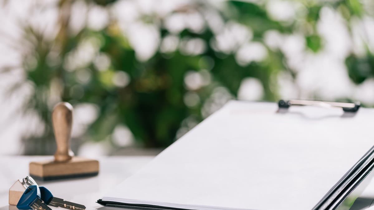 A set of house keys beside a clipboard of papers on a white table, a wooden stamp and green plants behind