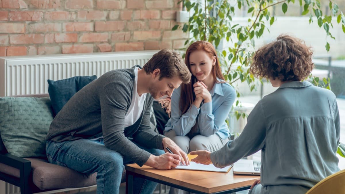 A young couple on a sofa signing papers at a low table while a counselor across from them points to a line