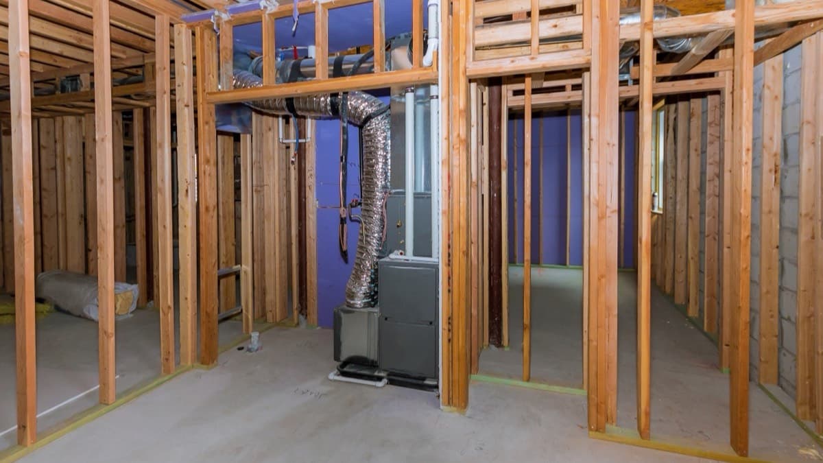 A gas furnace in a framed basement, with a flexible duct rising from its top and white vent pipes along the joists