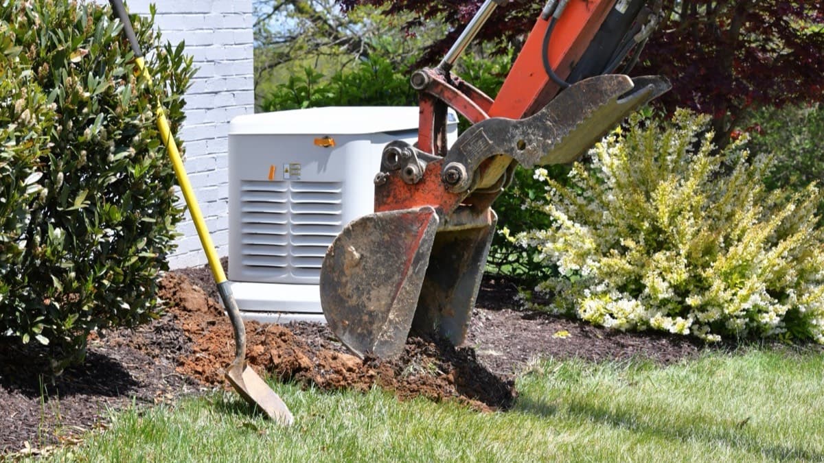 A standby generator beside a brick house while an excavator digs the trench for its lines