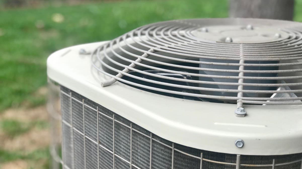 The fan grille on top of a central air conditioning condenser outside a house, with a summer lawn behind it
