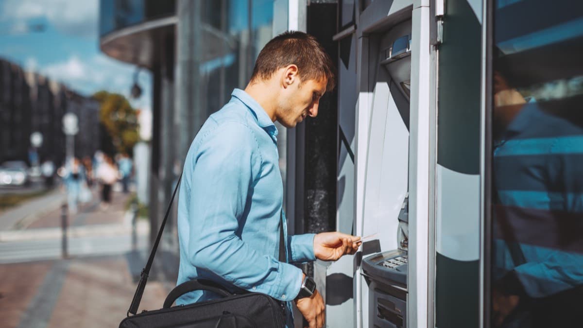 A man in a blue shirt putting his card into a bank's ATM on a sunny city street