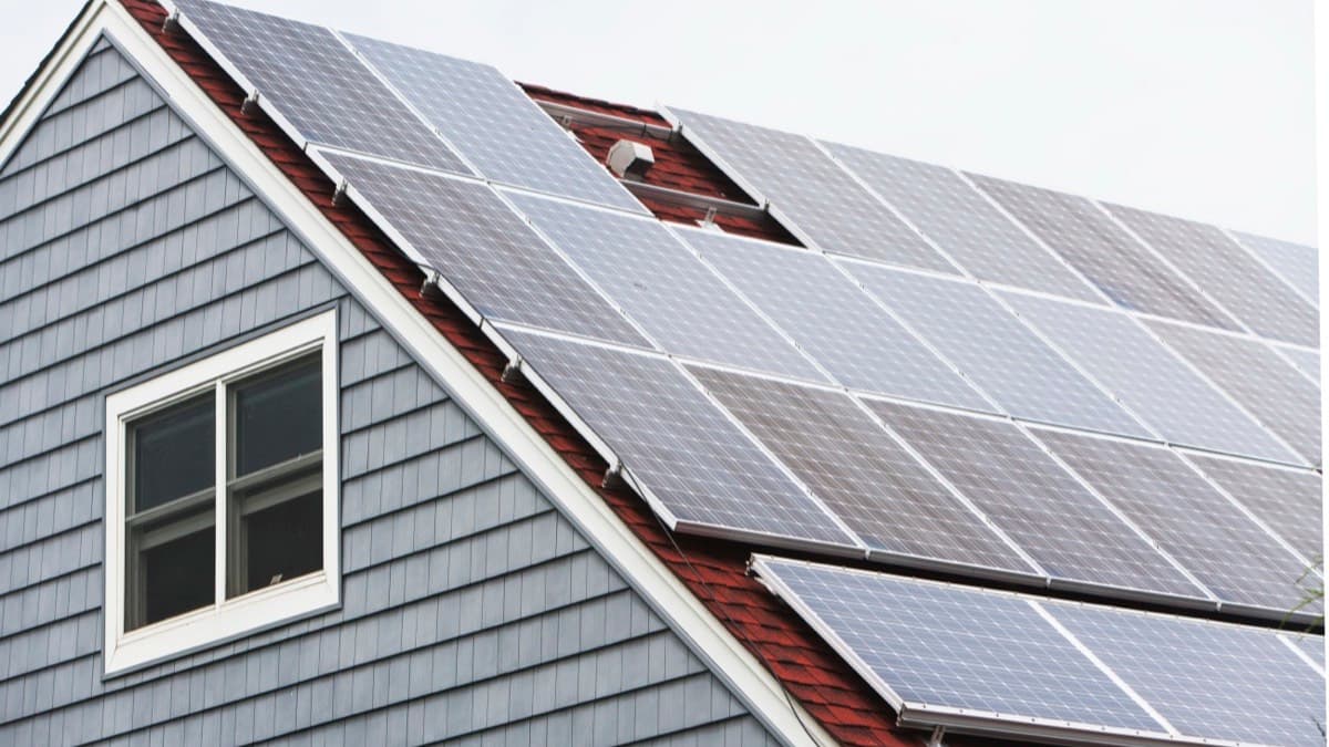 Rows of solar panels mounted across the shingled roof of a gray-sided house under a clear sky
