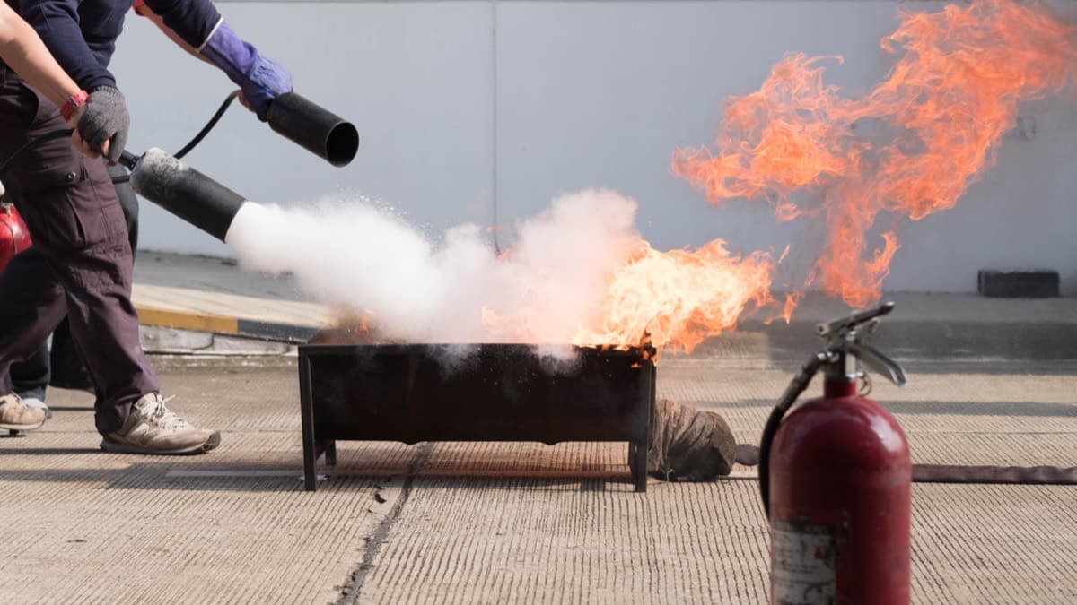 A person in gloves spraying a fire extinguisher at the base of a pan fire during a training demonstration, with a second extinguisher standing by