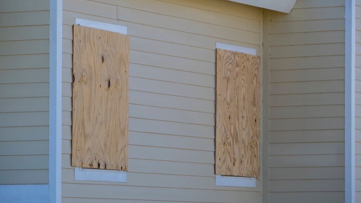 Two windows on a pale yellow sided house covered with plywood sheets ahead of a storm