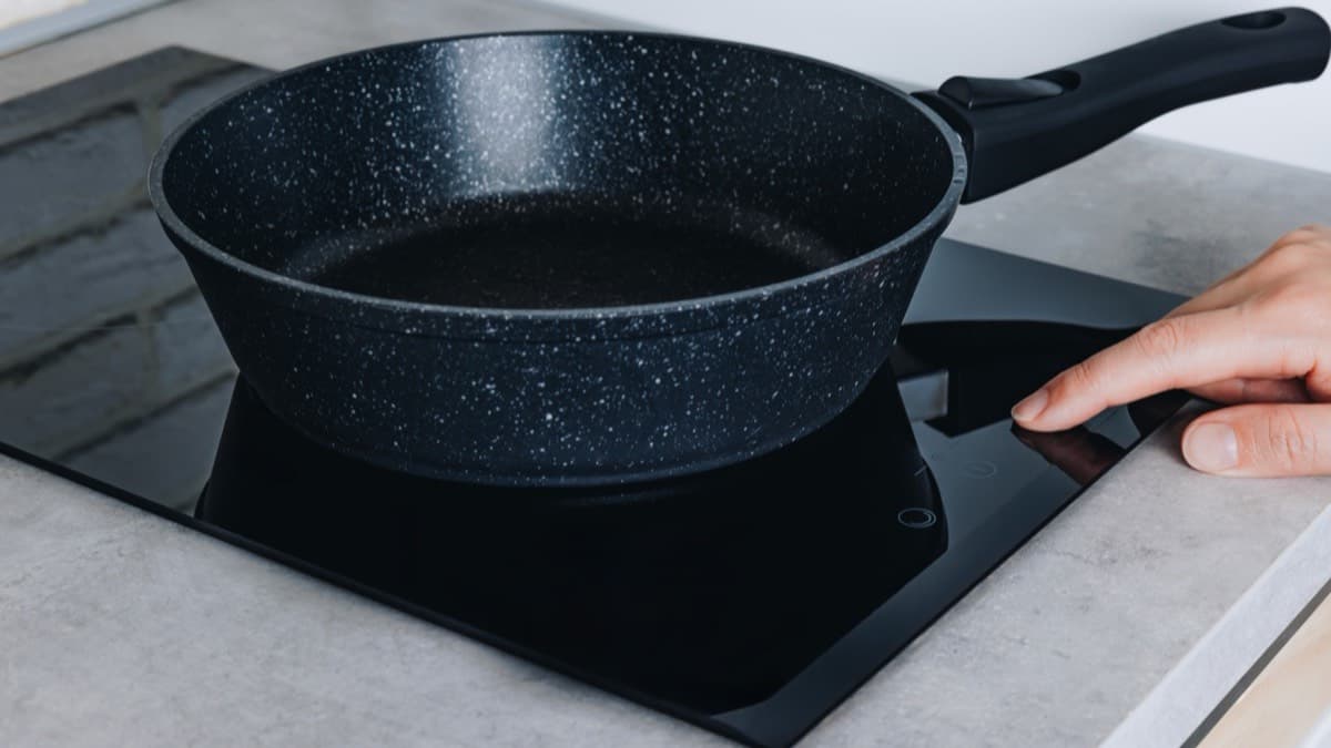 A black frying pan on a glass induction cooktop with a finger touching the control panel