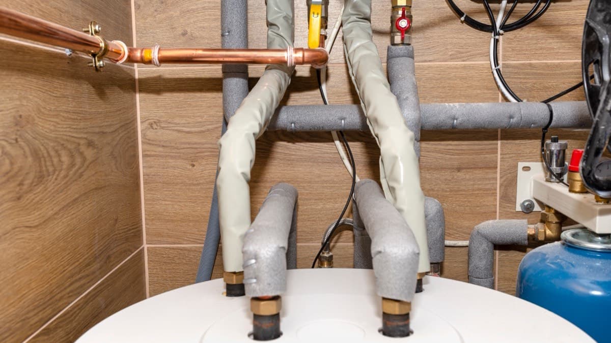 Gray foam insulation sleeves over the copper pipes rising from the top of a water heater, beside a blue expansion tank