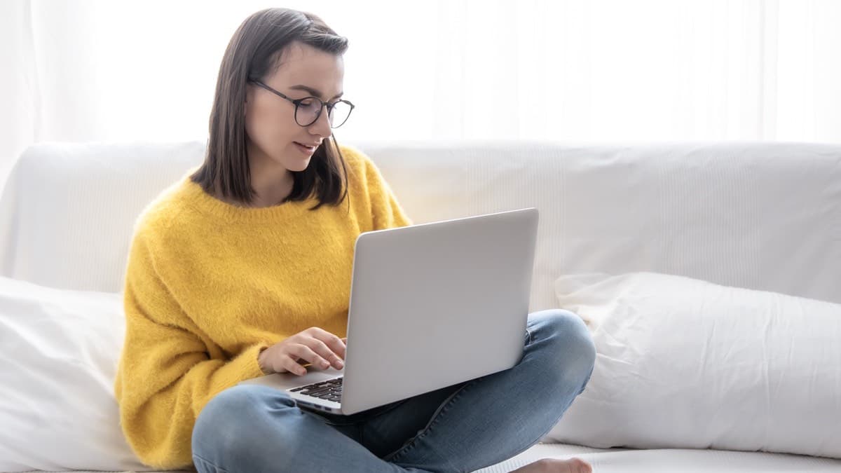 A woman in a yellow sweater working on a laptop on a sofa