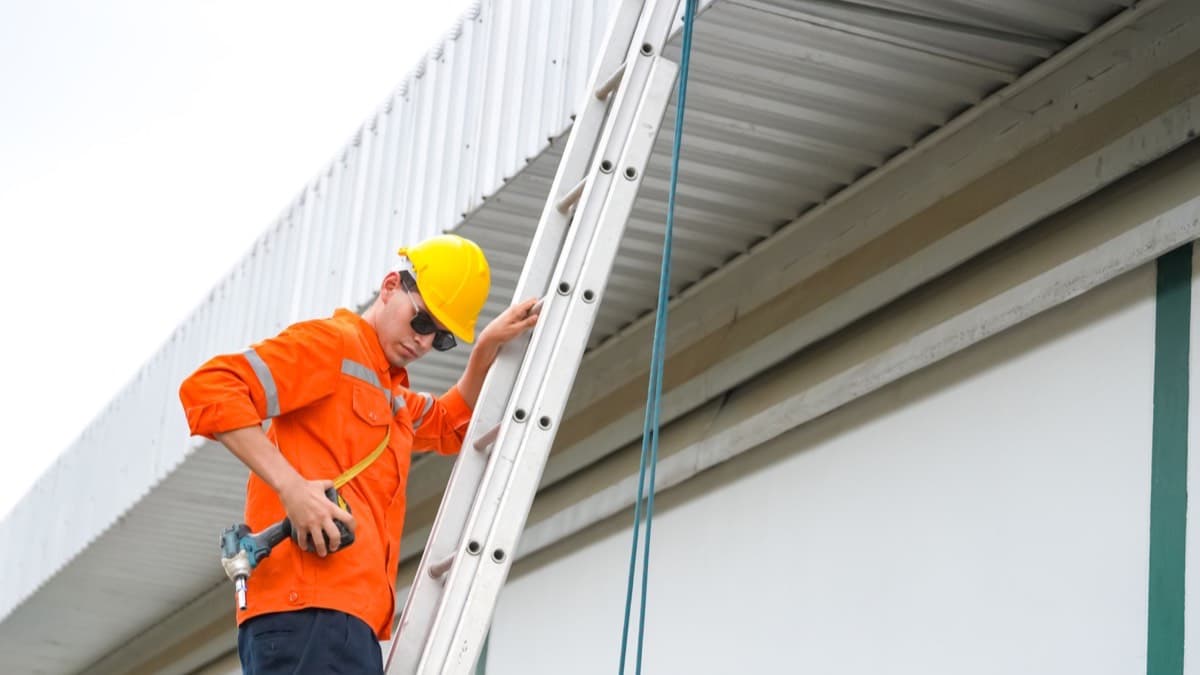A construction worker in a hard hat climbing an extension ladder to a roof edge