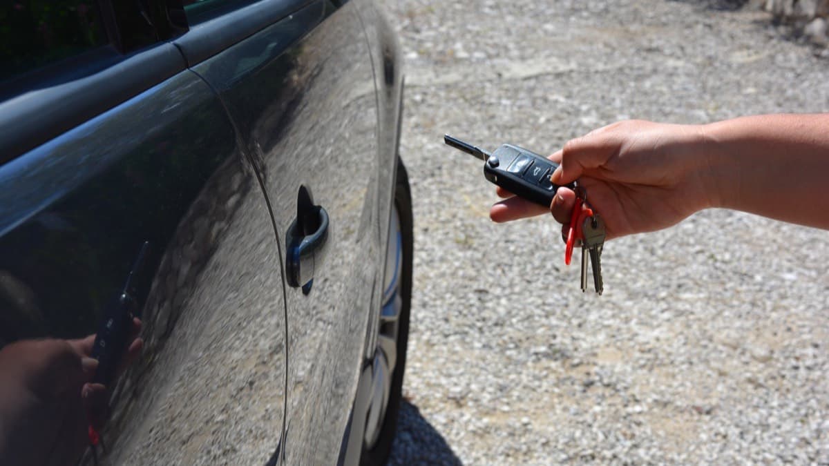 A hand pointing a key fob at a parked car on a gravel driveway